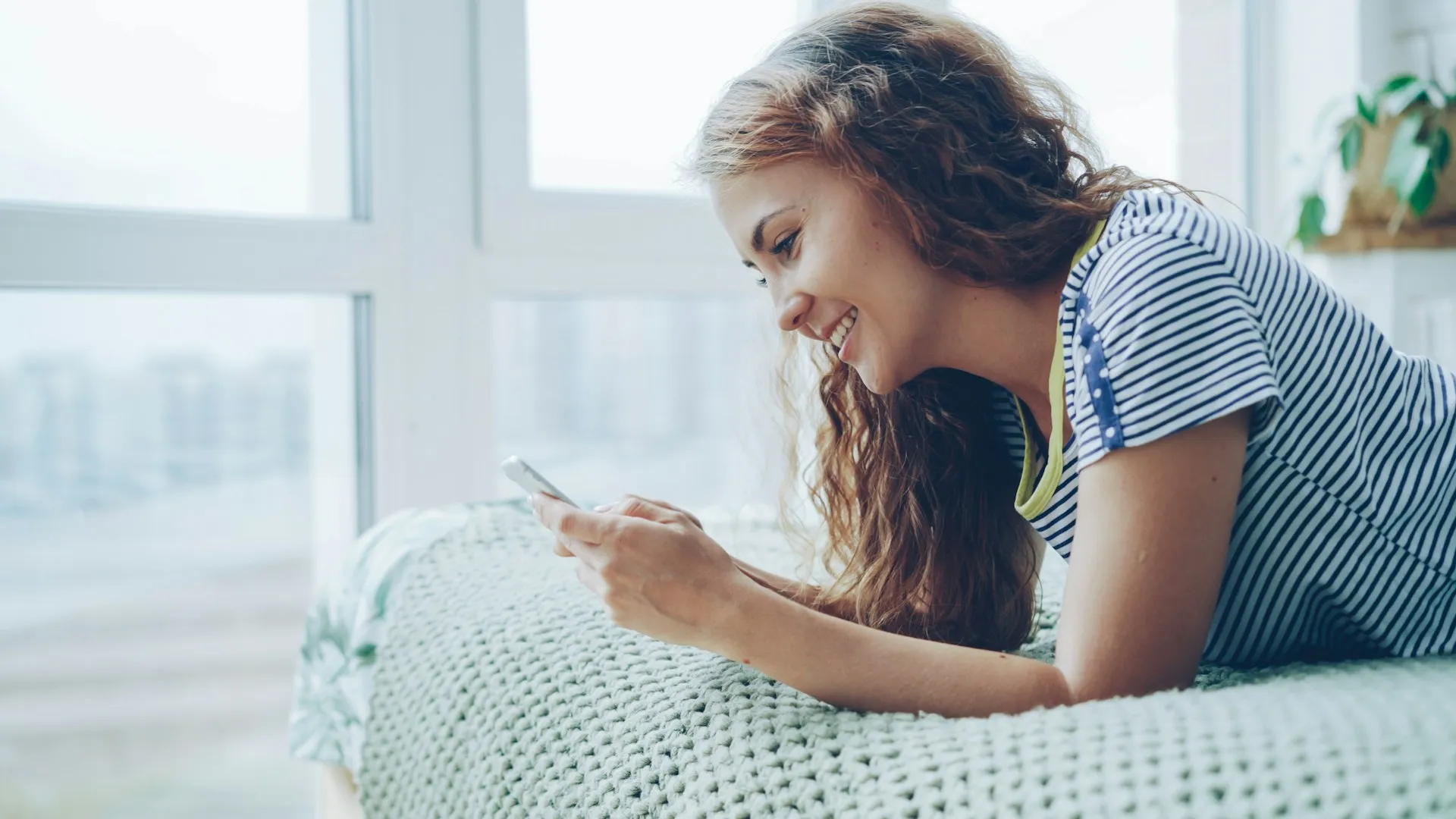 A happy woman lying on bed at home using her smartphone.